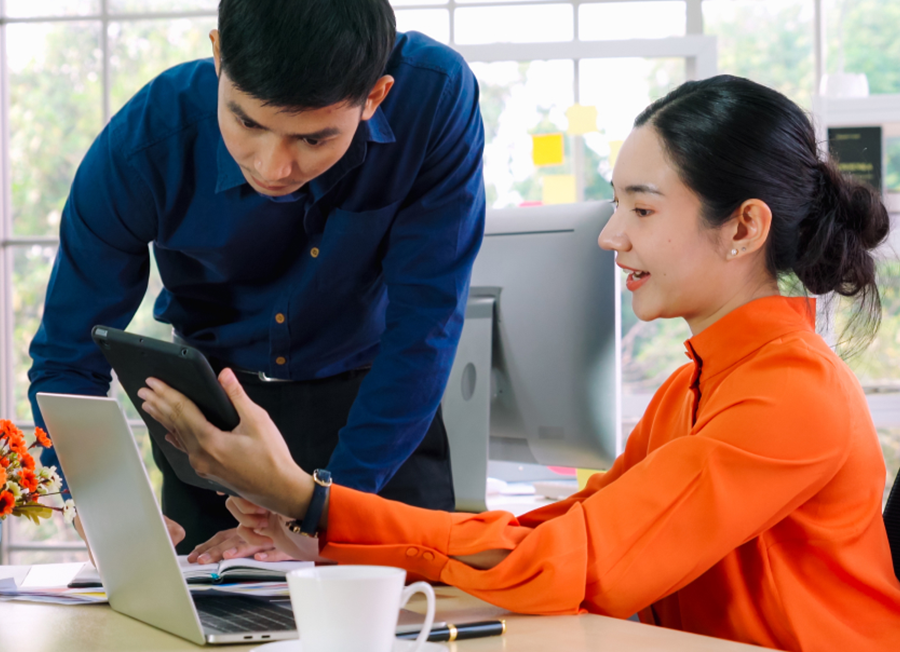 man leaning down on desk next a to a woman using laptop and showing a tablet screen