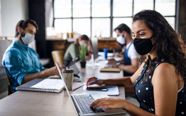 woman wearing mask sitting at desk on laptop with others